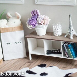May include: A white shelf unit with three shelves, a white base, and a brown top. The top shelf has a small white pot with purple and white flowers, a white Eiffel Tower figurine, and a glass jar with white stones. The middle shelf has a black sign that says "Home". The bottom shelf has a stack of books. The shelf is sitting on a black and white cowhide rug.