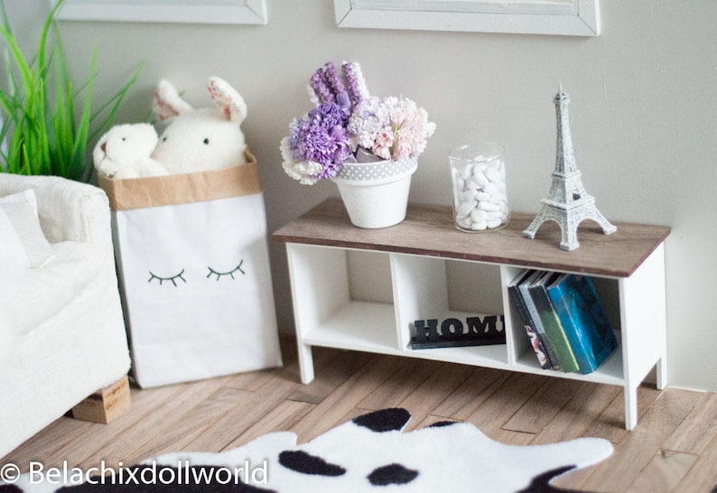 May include: A white shelf unit with three shelves, a white base, and a brown top. The top shelf has a small white pot with purple and white flowers, a white Eiffel Tower figurine, and a glass jar with white stones. The middle shelf has a black sign that says "Home". The bottom shelf has a stack of books. The shelf is sitting on a black and white cowhide rug.