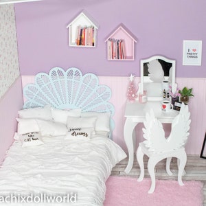 May include: A white bed with a blue and white headboard, white pillows with black text, and a white blanket. The bed is in a room with a white vanity with a mirror, a white chair with wings, and a white lamp. The walls are lavender and the floor is light wood.
