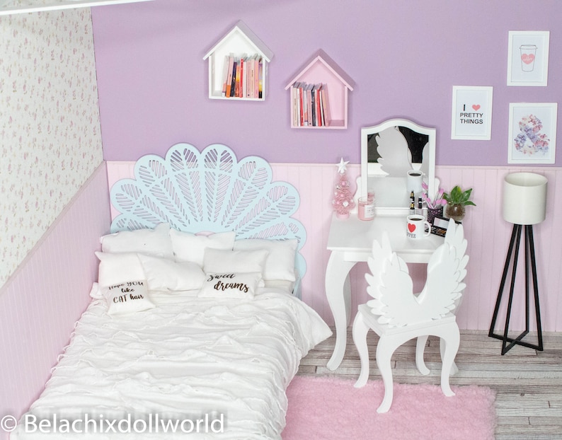 May include: A white bed with a blue and white headboard, white pillows with black text, and a white blanket. The bed is in a room with a white vanity with a mirror, a white chair with wings, and a white lamp. The walls are lavender and the floor is light wood.