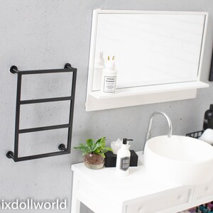 May include: A white bathroom vanity with a white porcelain sink and a black metal towel rack. The vanity has a white countertop with a black soap dispenser, a small potted plant, and a white tissue box. There is a black framed print on the wall that says "Beauty is an attitude".