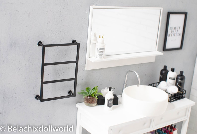May include: A white bathroom vanity with a white porcelain sink and a black metal towel rack. The vanity has a white countertop with a black soap dispenser, a small potted plant, and a white tissue box. There is a black framed print on the wall that says "Beauty is an attitude".