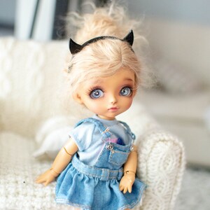 May include: A small doll with blonde hair and blue eyes wearing a denim overall dress and a black headband with devil horns. The doll is sitting on a white knitted surface.