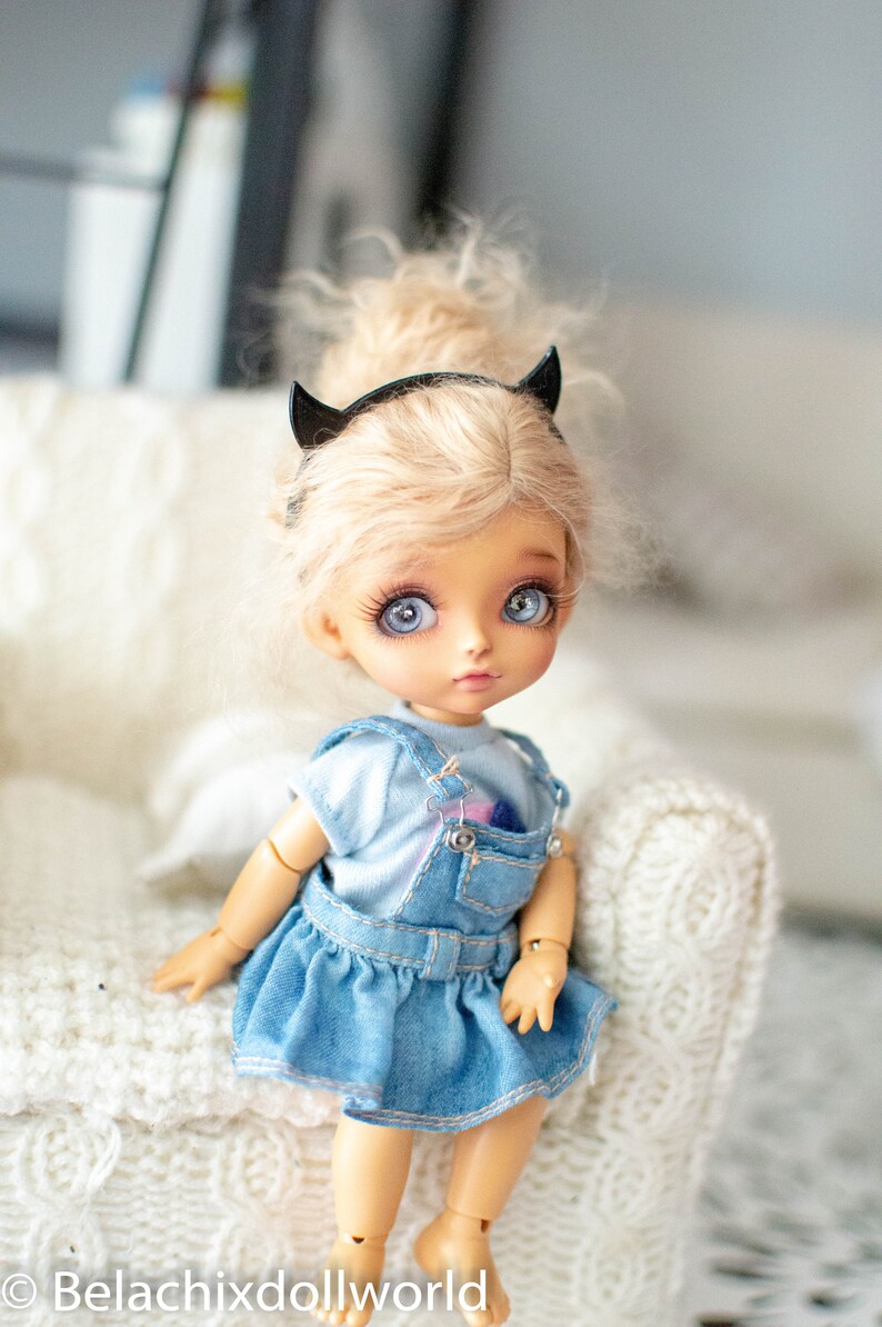 May include: A small doll with blonde hair and blue eyes wearing a denim overall dress and a black headband with devil horns. The doll is sitting on a white knitted surface.