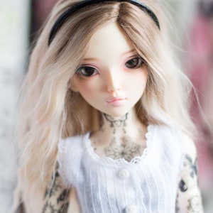 May include: A doll with long, blonde hair wearing a white dress with lace trim and a black headband. The doll has a delicate face with blue eyes and a tattoo on her chest.