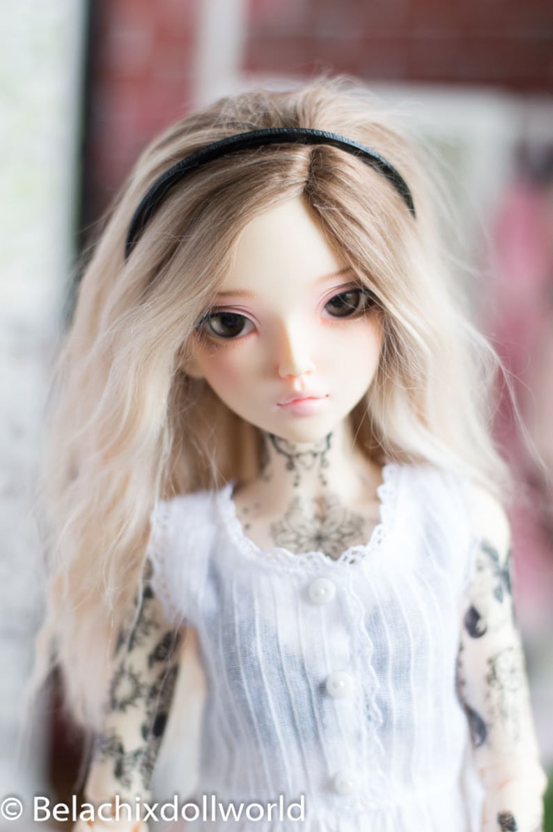 May include: A doll with long, blonde hair wearing a white dress with lace trim and a black headband. The doll has a delicate face with blue eyes and a tattoo on her chest.