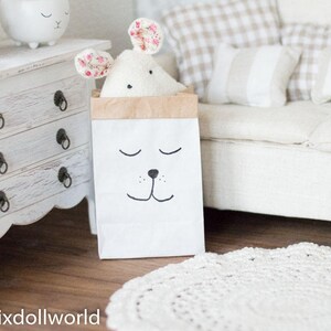 May include: A white stuffed animal with pink floral ears is inside a brown paper bag with a black and white cartoon face. The bag is sitting on a wooden floor in front of a white couch with beige and white pillows.