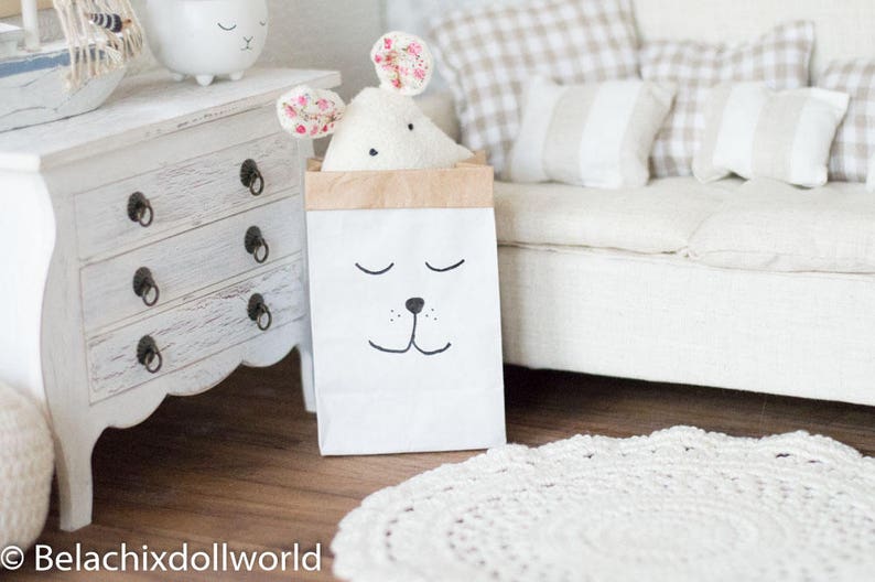 May include: A white stuffed animal with pink floral ears is inside a brown paper bag with a black and white cartoon face. The bag is sitting on a wooden floor in front of a white couch with beige and white pillows.