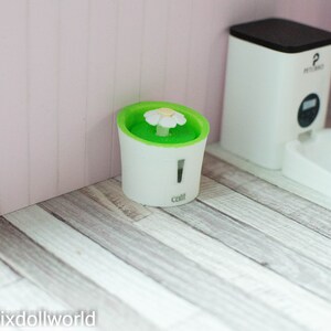 May include: A white pet fountain with a green water basin and a white flower on top. The fountain is labeled "catit" and is sitting on a light wood floor.