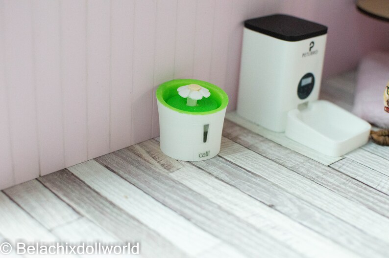 May include: A white pet fountain with a green water basin and a white flower on top. The fountain is labeled "catit" and is sitting on a light wood floor.