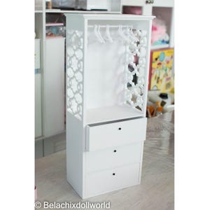 May include: A white doll-sized wardrobe with a decorative openwork design on the side. The wardrobe has a top shelf with a rod for hanging clothes and three drawers. Several white hangers are inside the wardrobe.