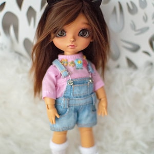 May include: A small doll with brown hair wearing a pink shirt and blue overalls. The doll has brown eyes and is wearing a black headband with devil horns. The doll is standing on a white fluffy surface.