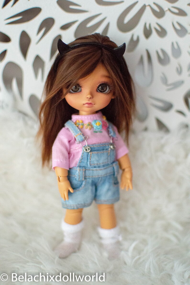 May include: A small doll with brown hair wearing a pink shirt and blue overalls. The doll has brown eyes and is wearing a black headband with devil horns. The doll is standing on a white fluffy surface.