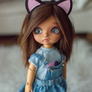 May include: A doll with brown hair and large blue eyes wearing a denim dress and a light blue shirt. The doll has a black headband with pink cat ears. The doll's outfit includes a denim overall dress with a pocket.