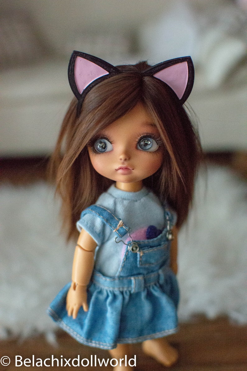 May include: A doll with brown hair and large blue eyes wearing a denim dress and a light blue shirt. The doll has a black headband with pink cat ears. The doll's outfit includes a denim overall dress with a pocket.
