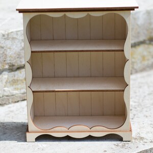 Pu&ograve; includere: Un piccolo scaffale in legno bianco con tre ripiani. I ripiani hanno un bordo a festoni e l'unit&agrave; ha un design semplice e rustico.