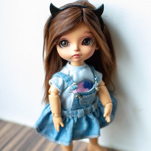 May include: A small doll with brown hair wearing a blue denim overall dress and a black headband with horns. The doll has brown eyes and a light skin tone.