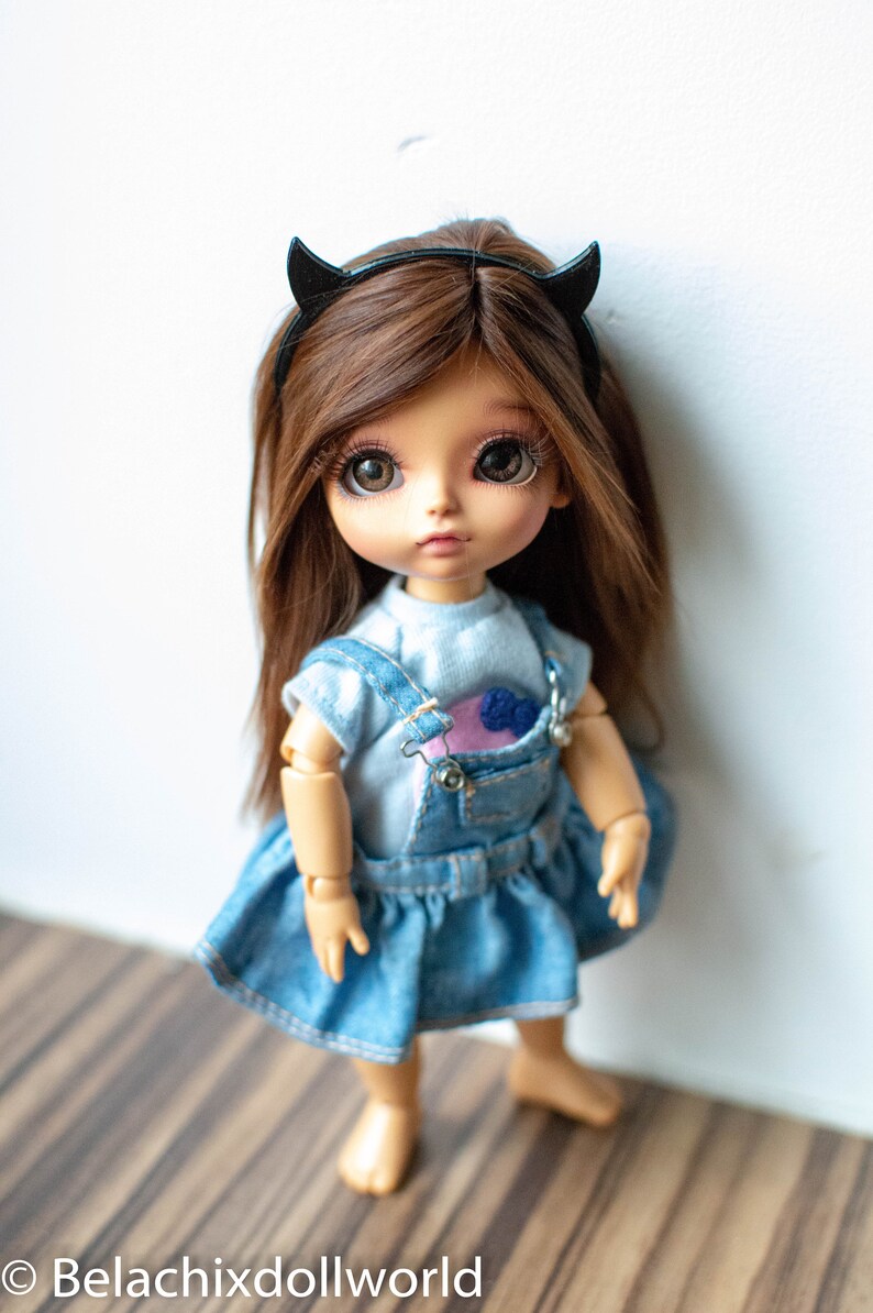 May include: A small doll with brown hair wearing a blue denim overall dress and a black headband with horns. The doll has brown eyes and a light skin tone.
