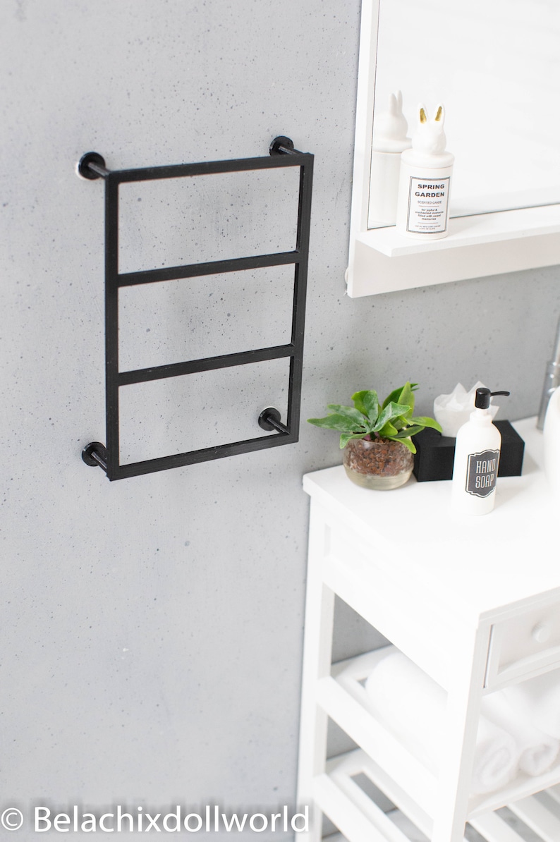 May include: Black metal towel rack with three bars mounted on a white wall. A white shelf with a bottle of hand soap and a small plant is visible in the background.
