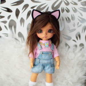 May include: A doll with brown hair and large eyes wearing a pink shirt, denim overalls, and white socks. The doll has a cat ear headband. The background is a white surface with a floral pattern.