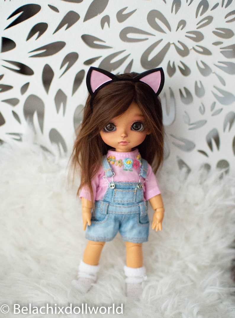 May include: A doll with brown hair and large eyes wearing a pink shirt, denim overalls, and white socks. The doll has a cat ear headband. The background is a white surface with a floral pattern.