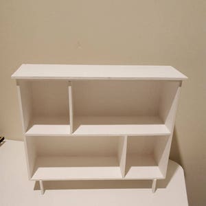 May include: White wooden shelf unit with five compartments. The shelf has a simple design and is perfect for displaying small items.