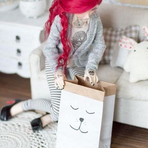 May include: A red-haired doll wearing a grey cardigan and striped pants is holding a brown paper bag with a white sleeping animal face drawn on it.