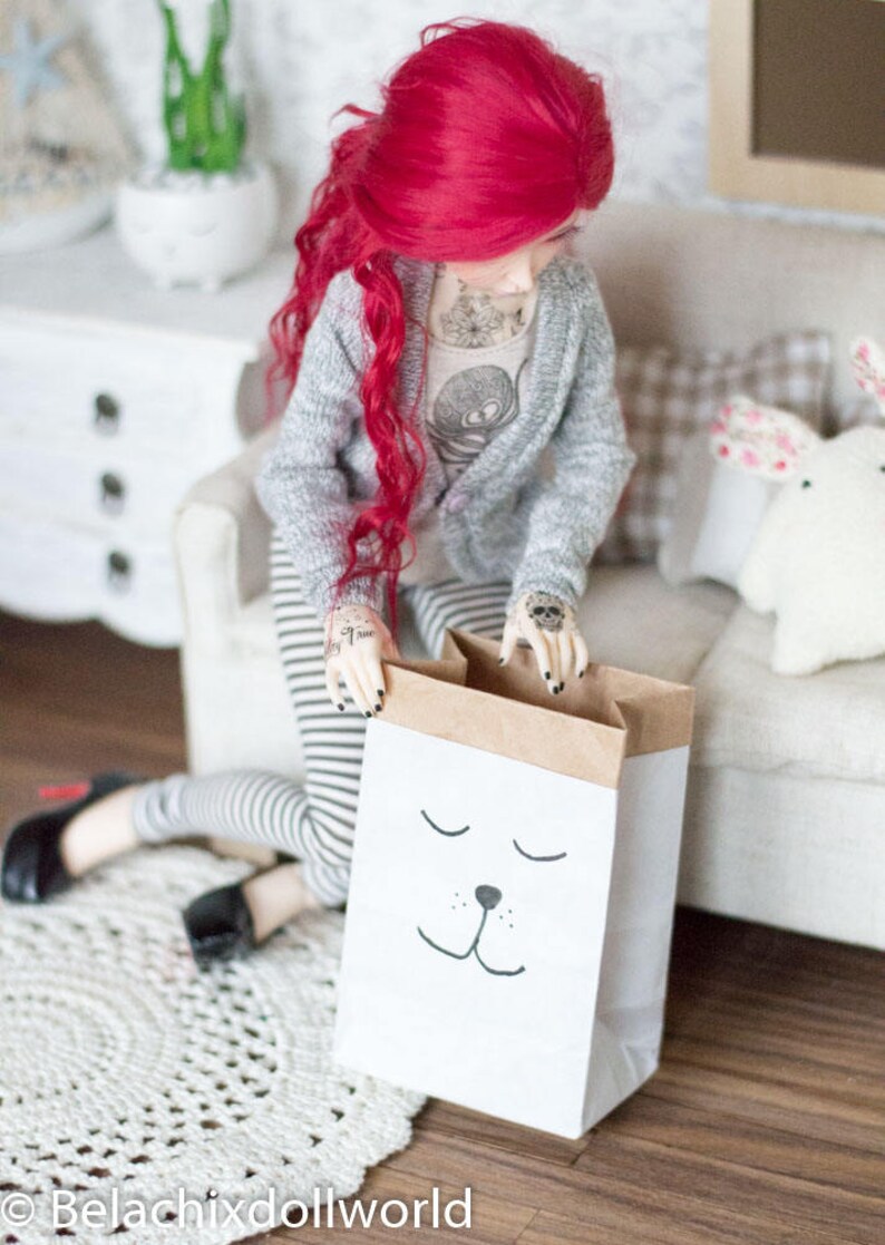 May include: A red-haired doll wearing a grey cardigan and striped pants is holding a brown paper bag with a white sleeping animal face drawn on it.