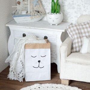 May include: A white paper bag with a brown top and a black cartoon face with closed eyes and a nose. The bag is sitting on a white wooden dresser with a white crocheted blanket and a white ceramic planter with a green succulent plant.