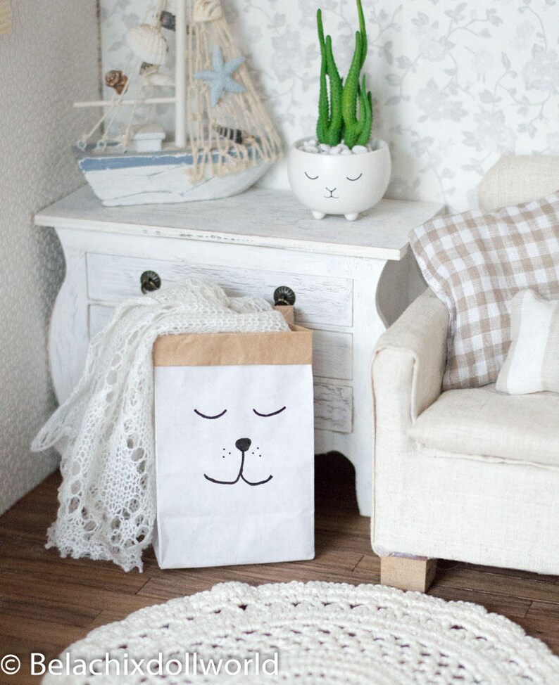 May include: A white paper bag with a brown top and a black cartoon face with closed eyes and a nose. The bag is sitting on a white wooden dresser with a white crocheted blanket and a white ceramic planter with a green succulent plant.