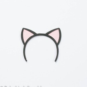 May include: A black headband with pink cat ears. The ears are outlined in black and have a pink interior. The headband is designed to be worn on the head and is a cute accessory.