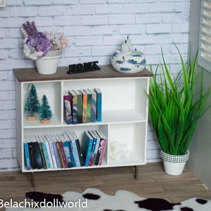 May include: White bookshelf with three shelves, filled with books and small potted plants. The bookshelf is against a white brick wall. The top shelf has a decorative bowl with candles and the word "Home" in black letters.