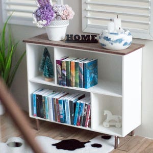 May include: A white bookshelf with four shelves, filled with books and a small artificial Christmas tree. The bookshelf is on a wooden surface with a white cowhide rug in front of it. There is a white ceramic planter with purple flowers on the shelf above the bookshelf. The word "HOME" is written in black letters on the shelf above the bookshelf.