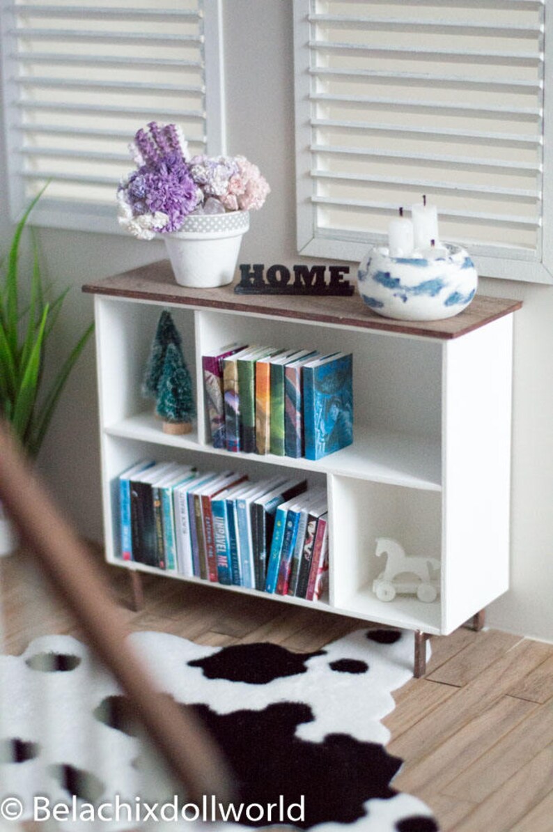 May include: A white bookshelf with four shelves, filled with books and a small artificial Christmas tree. The bookshelf is on a wooden surface with a white cowhide rug in front of it. There is a white ceramic planter with purple flowers on the shelf above the bookshelf. The word "HOME" is written in black letters on the shelf above the bookshelf.