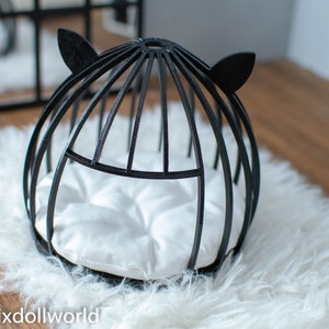 May include: Black cat bed with a white cushion inside. The bed is made of wood and has a round shape with cat ears on the top.
