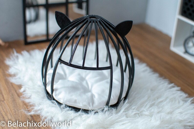 May include: Black cat bed with a white cushion inside. The bed is made of wood and has a round shape with cat ears on the top.