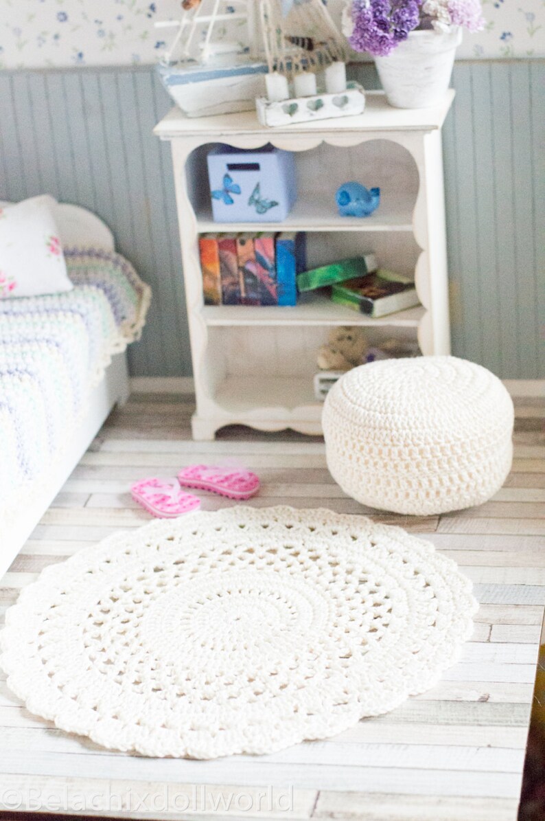 Pu&ograve; includere: Un tappeto rotondo in crochet bianco con un motivo delicato. Il tappeto &egrave; su un pavimento in legno chiaro con un pouf in crochet bianco e infradito rosa.