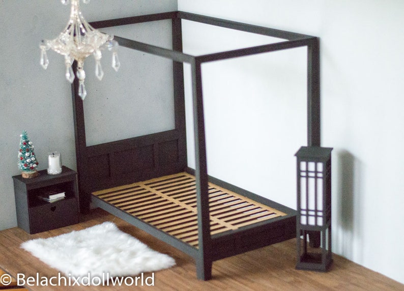 Puede incluir: Una cama con dosel negro con un colch&oacute;n de listones de madera y una alfombra blanca y esponjosa. Una peque&ntilde;a mesita de noche negra con una vela y un &aacute;rbol de Navidad en miniatura se encuentra junto a la cama. Una l&aacute;mpara de pie negra con una pantalla blanca se encuentra en la esquina.