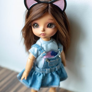 May include: A doll with brown hair and large eyes wearing a denim dress with a blue top and overalls. The doll has a cat ear headband. The doll is standing in front of a white wall. The text "Belachixdollworld" is at the bottom.