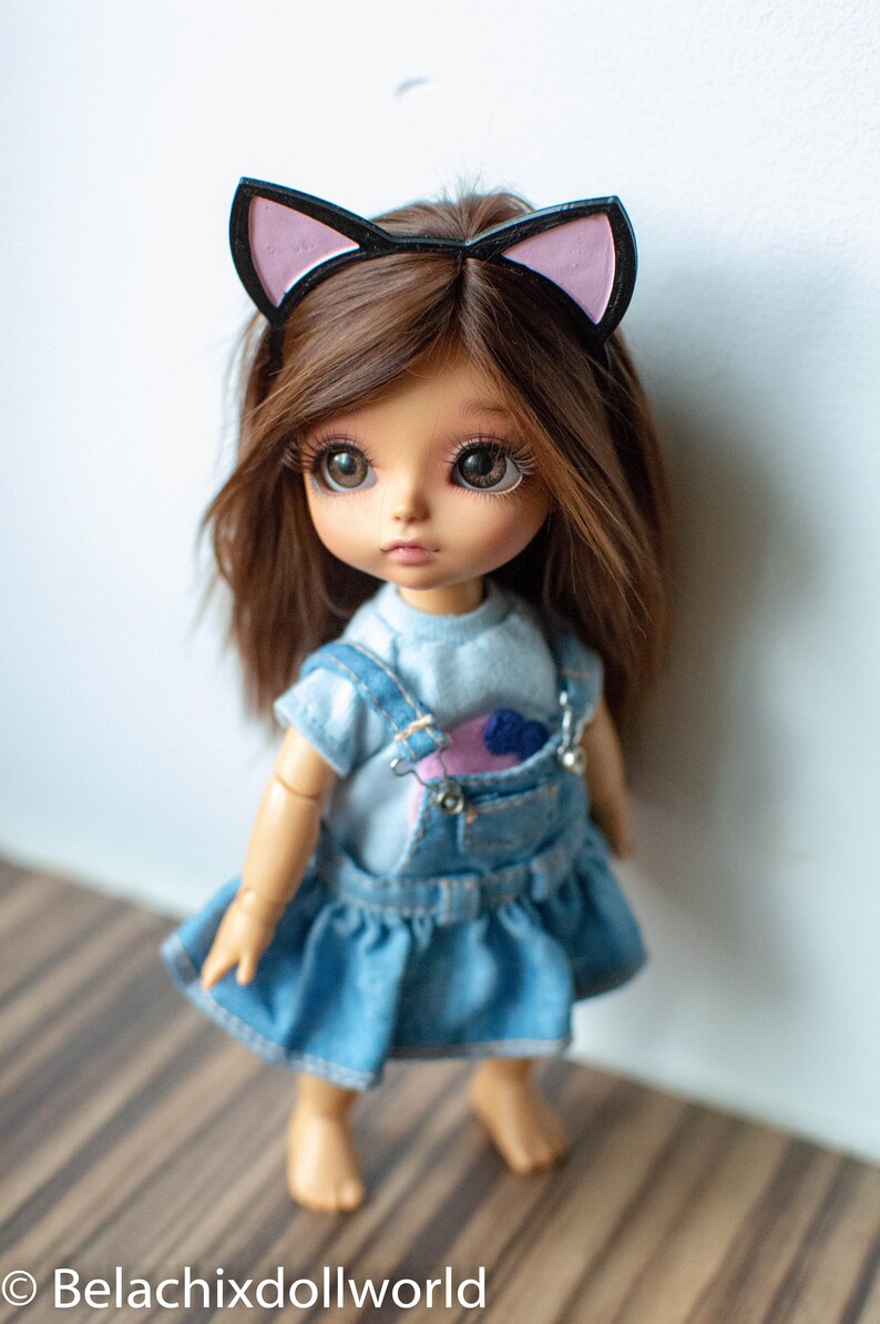 May include: A doll with brown hair and large eyes wearing a denim dress with a blue top and overalls. The doll has a cat ear headband. The doll is standing in front of a white wall. The text "Belachixdollworld" is at the bottom.