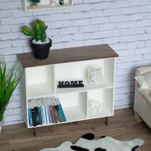 May include: White bookshelf with six compartments, featuring a cactus plant, books, a glass jar with white stones, and a black sign that reads "HOME". The bookshelf is on a wooden floor with a black and white cowhide rug.