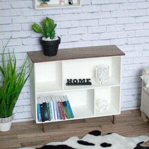 May include: White bookshelf with four shelves, a wooden top, and metal legs. The shelves are filled with books, a glass jar with white stones, and a black sign that says "HOME". A black and white cowhide rug is in front of the bookshelf.