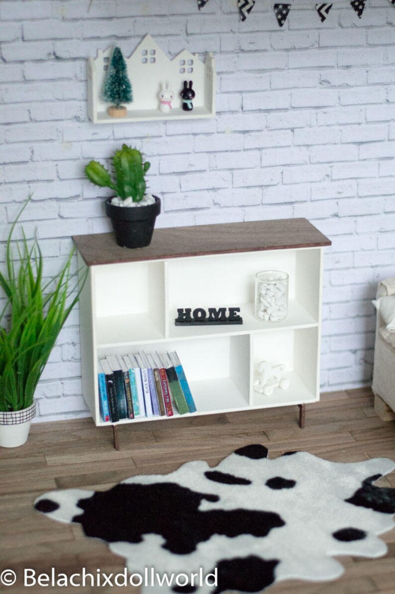 May include: White bookshelf with four shelves, a wooden top, and metal legs. The shelves are filled with books, a glass jar with white stones, and a black sign that says "HOME". A black and white cowhide rug is in front of the bookshelf.