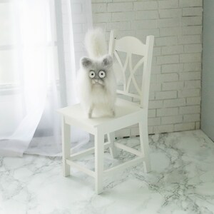 Peut inclure: Une chaise blanche avec un chat en feutre blanc et moelleux assis sur le si&egrave;ge. Le chat a de grands yeux noirs et un visage gris. La chaise et le chat sont devant un mur de briques blanches.