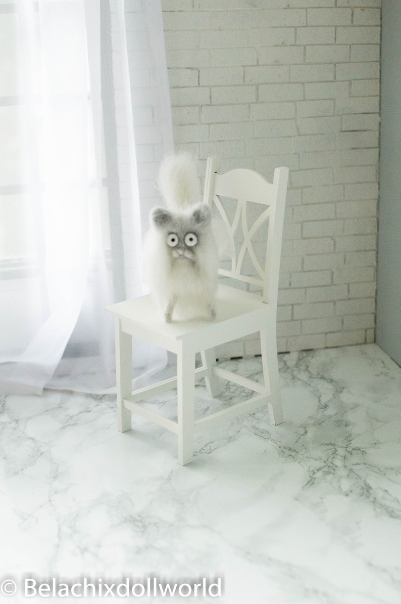 Peut inclure: Une chaise blanche avec un chat en feutre blanc et moelleux assis sur le si&egrave;ge. Le chat a de grands yeux noirs et un visage gris. La chaise et le chat sont devant un mur de briques blanches.