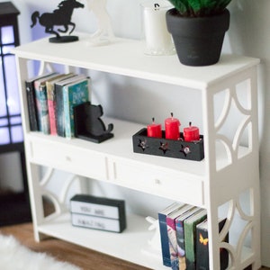 May include: White bookshelf with three shelves and two drawers. The top shelf has a black horse figurine, a white horse figurine, and a potted plant. The middle shelf has books, a black bookend, and a black tray with four red candles. The bottom shelf has books, a black sign that says "You Are Amazing", and a white rabbit figurine.