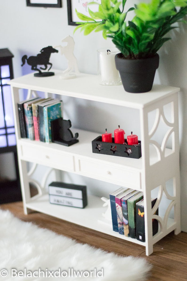 May include: White bookshelf with three shelves and two drawers. The top shelf has a black horse figurine, a white horse figurine, and a potted plant. The middle shelf has books, a black bookend, and a black tray with four red candles. The bottom shelf has books, a black sign that says "You Are Amazing", and a white rabbit figurine.