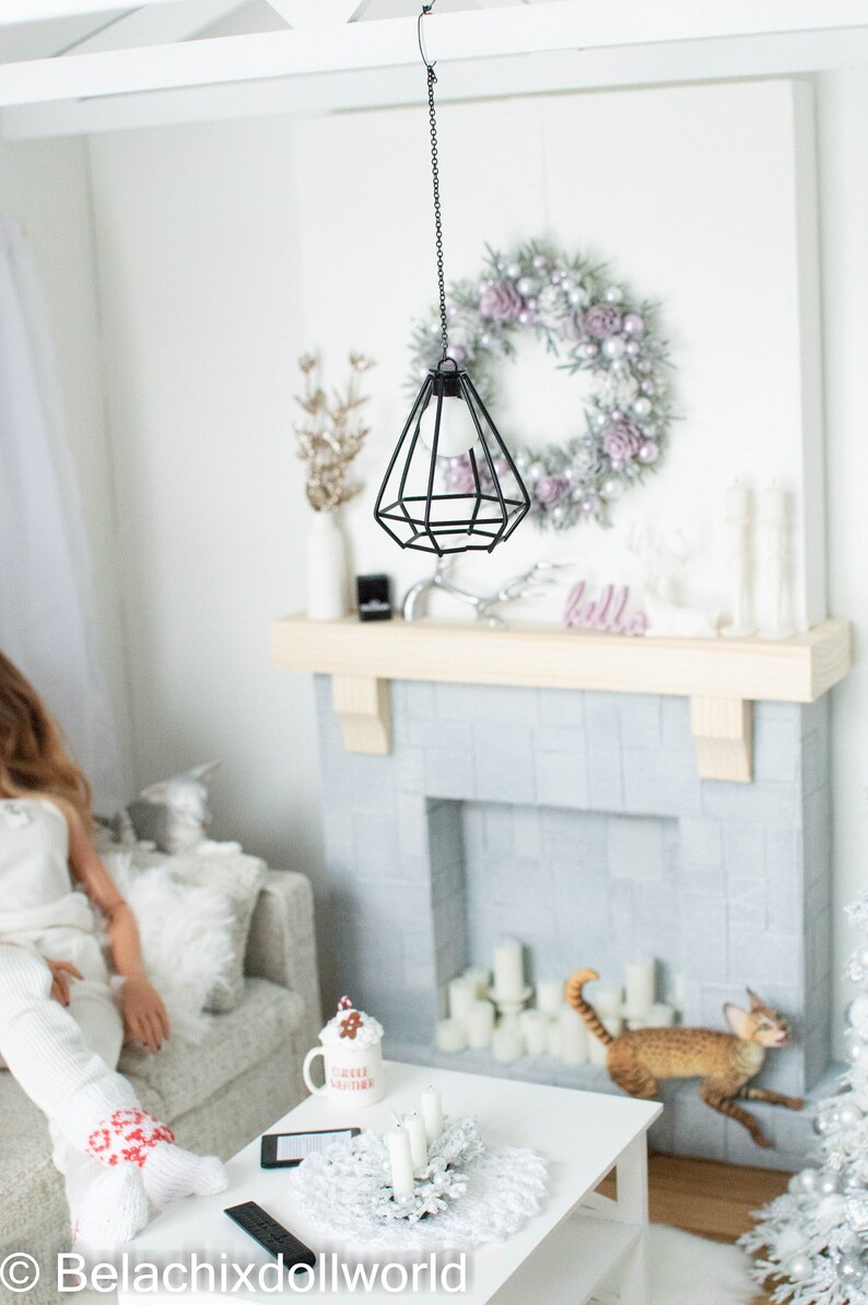 May include: A miniature dollhouse living room with a white fireplace, a white couch, a white coffee table, a white Christmas tree, a white mug with the text "Coffee Mother", a white wreath with silver and pink ornaments, and a black hanging light fixture.