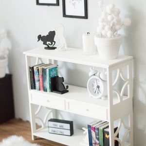 May include: White bookshelf with two shelves and two drawers. The top shelf has a white potted plant with white pom poms, a glass candle holder, and a black silhouette of a horse. The middle shelf has a set of books with black bookends shaped like a cat. A white alarm clock is on the right side of the shelf. The bottom shelf has a black sign that says "You are amazing" and another set of books.