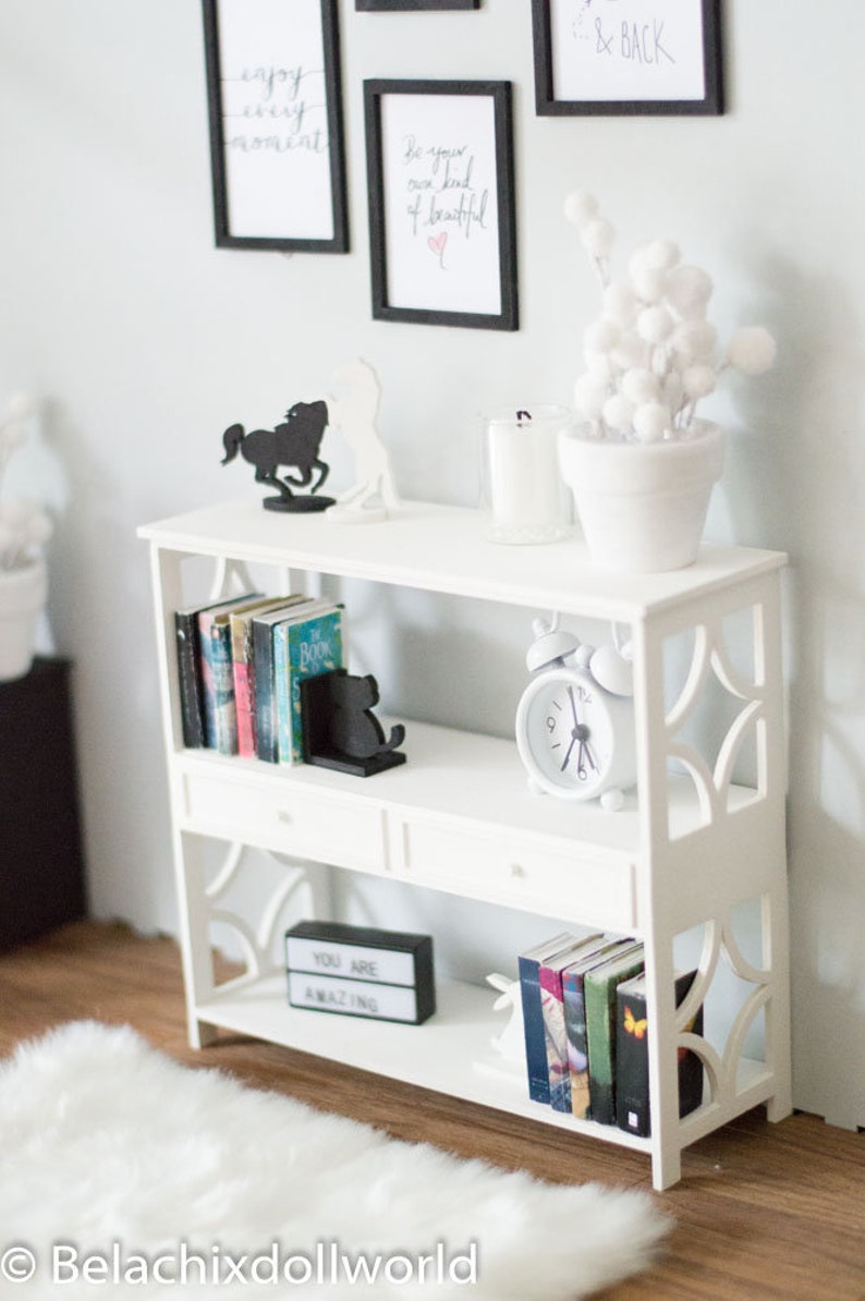May include: White bookshelf with two shelves and two drawers. The top shelf has a white potted plant with white pom poms, a glass candle holder, and a black silhouette of a horse. The middle shelf has a set of books with black bookends shaped like a cat. A white alarm clock is on the right side of the shelf. The bottom shelf has a black sign that says "You are amazing" and another set of books.
